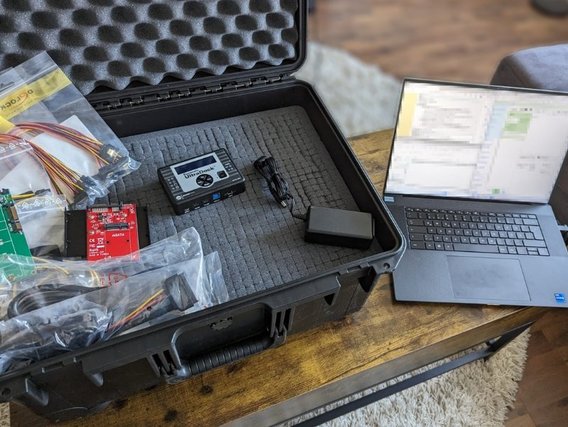 Ein Koffer mit elektronischen Bauteilen, darunter eine rote Platine und verschiedene Kabel, neben einem Laptop auf einem Holztisch.