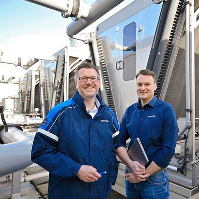 Zwei Personen stehen vor einer Reihe großer technischer Anlagen mit Rohrleitungen und Wärmetauschern in einer industriellen Umgebung.