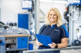 TÜV NORD Mitarbeiterin mit Klemmbrett in einer Werkstatt, im Hintergrund Werkstattausrüstung und ein Auto.