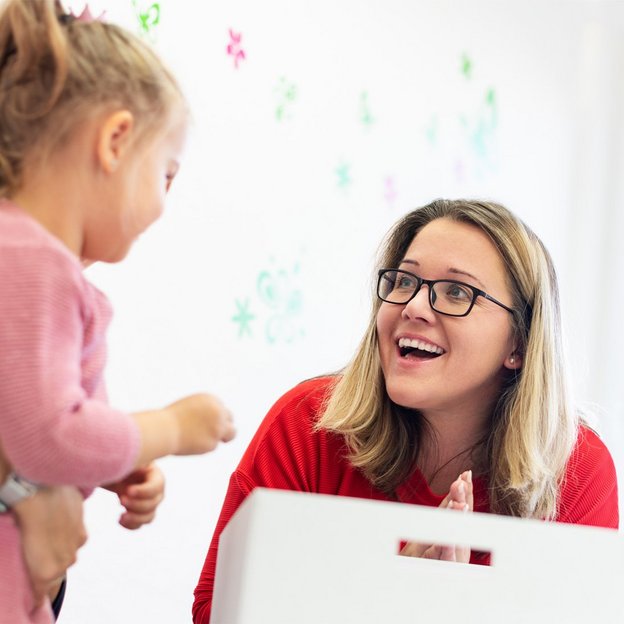 Eine Frau in einem roten Pullover interagiert mit einem kleinen Mädchen, das von einer anderen Person gehalten wird. Der Raum ist mit bunten Wanddekorationen versehen. Das Bild zeigt eine Coaching-Situation mit Fokus auf Kinderbetreuung.