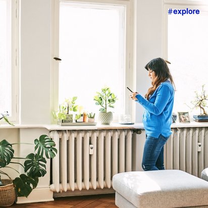 Eine Frau in blauem Oberteil und Jeans steht an einem Fenster mit vielen Zimmerpflanzen in einem hellen Wohnzimmer mit weißen Wänden.