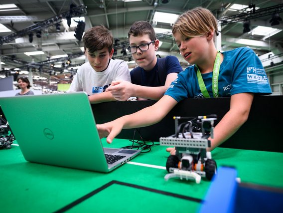 Drei Jungen arbeiten gemeinsam an einem Laptop mit grünem Gehäuse. Neben dem Computer steht ein kleiner Roboter auf einer grünen Matte. Die Jungen tragen verschiedenfarbige T-Shirts und diskutieren über die Programmierung.