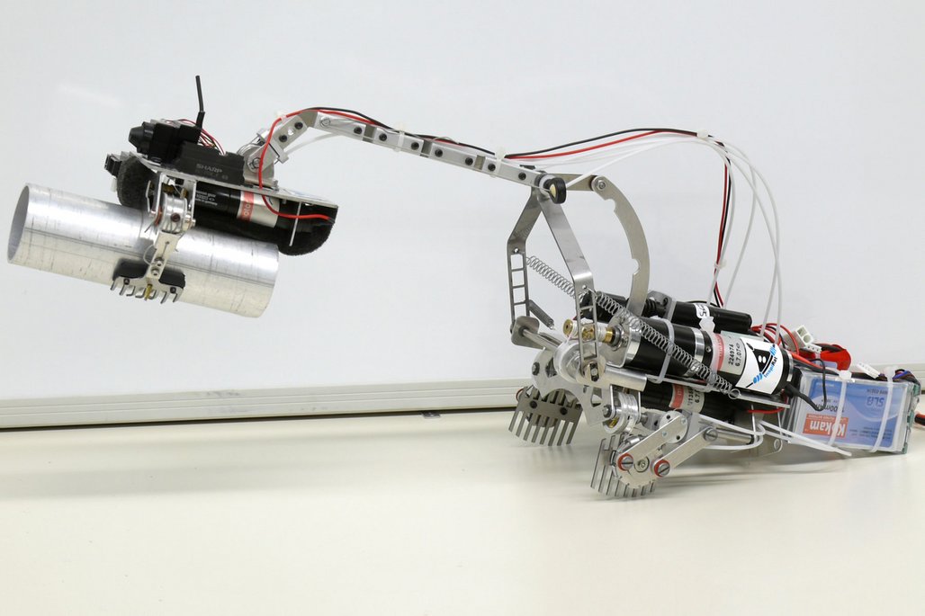 Ein selbstgebauter Roboterarm aus Metallteilen, Motoren und Elektronik. Er besteht aus mehreren Gelenken mit schwarzen Sharp-Motoren, roten und weißen Kabeln und einer metallischen Greifvorrichtung auf einer weißen Oberfläche.