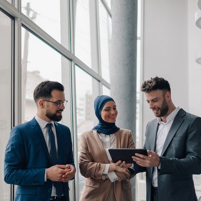Drei Personen in Business‑Kleidung stehen gemeinsam vor großen Fenstern und besprechen Inhalte auf einem Tablet.