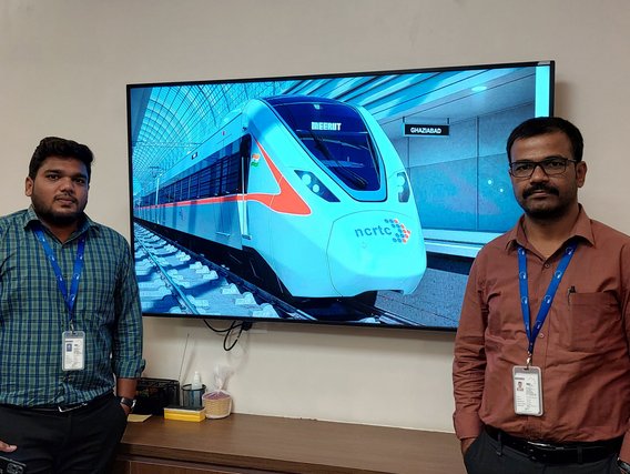 Sathish Kular Loganatahn (links) und Venkatesh Moode (rechts) stehen neben einem Bildschirm, der einen modernen Hochgeschwindigkeitszug (NCRTC) zeigt mit Anzeigen für Meerut und Ghaziabad.
