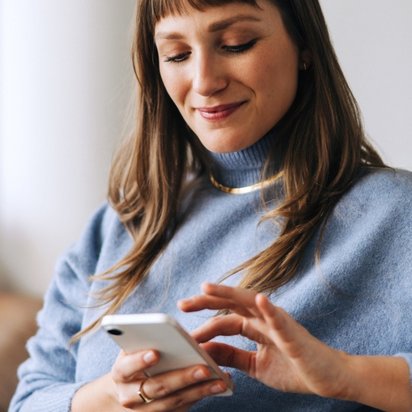 Eine Frau in blauem Pullover sitzt zu Hause auf einem Sofa und lächelt entspannt, während sie ihr Smartphone bedient.