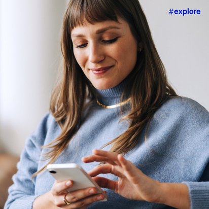 Eine Frau in blauem Pullover sitzt zu Hause auf einem Sofa und lächelt entspannt, während sie ihr Smartphone bedient.