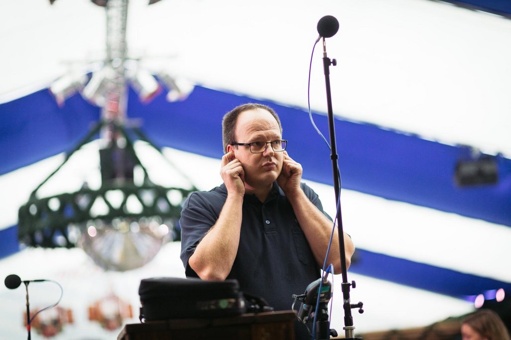 Ein Tontechniker unter einem blau-weißen Veranstaltungszelt prüft mit den Händen an den Ohren die Akustik neben einem aufgestellten Messmikrofon.