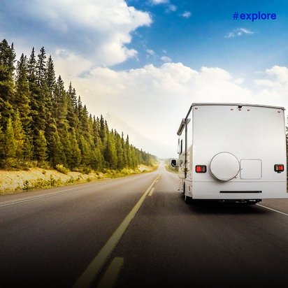 Ein weißes Wohnmobil fährt auf einer zweispurigen Straße durch eine bewaldete Landschaft mit Nadelbäumen. Der Himmel zeigt eine Mischung aus Wolken und blauem Himmel, während die Straße schnurgerade in die Ferne führt.