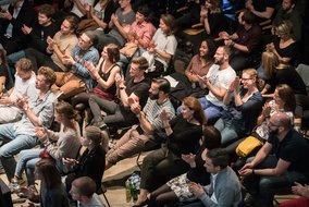 Eine applaudierende Publikumsgruppe in einem dunklen Raum, die in Sitzreihen angeordnet ist.