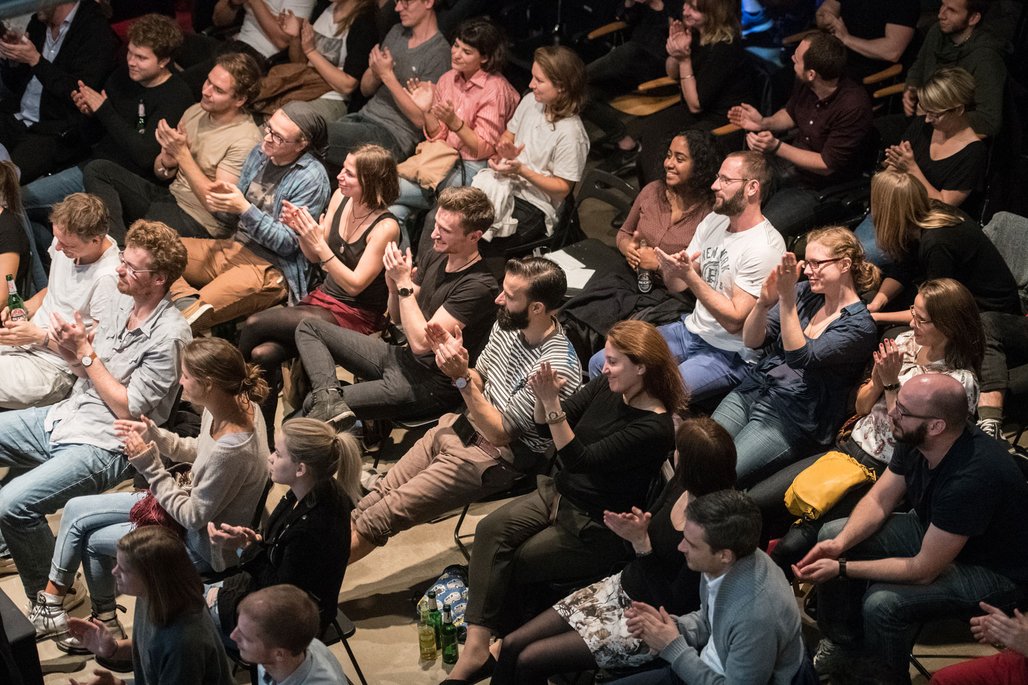 Eine applaudierende Publikumsgruppe in einem dunklen Raum, die in Sitzreihen angeordnet ist.