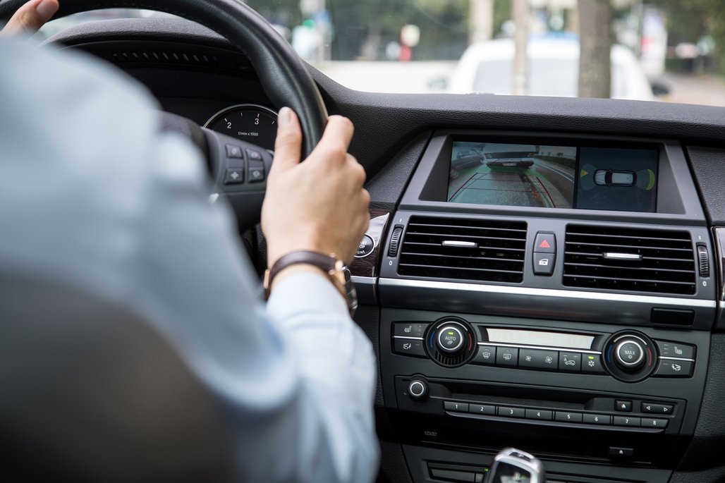 Ein Fahrer hält das schwarze Lenkrad eines Autos, am Handgelenk trägt er eine dunkle Uhr. Auf dem zentralen Display des Armaturenbretts ist die Rückfahrkamera-Ansicht mit grünen Führungslinien zu sehen, darunter befinden sich Klimaanlage und weitere Bedienelemente.
