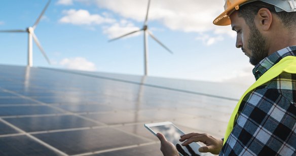 Techniker überprüft mit Tablet eine Photovoltaikanlage, im Hintergrund stehen Windräder.
