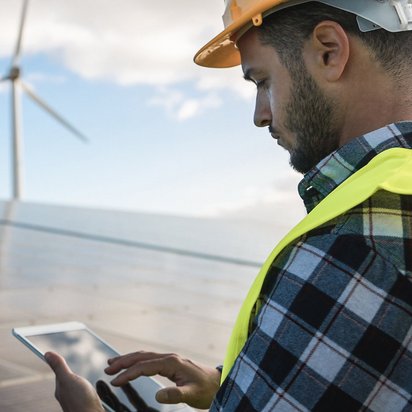 Techniker überprüft mit Tablet eine Photovoltaikanlage, im Hintergrund stehen Windräder.
