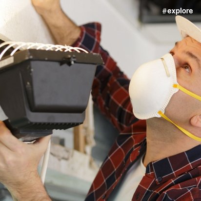 Ein Techniker mit Schutzhelm und Atemschutzmaske installiert oder prüft ein elektronisches Gerät an der Decke.