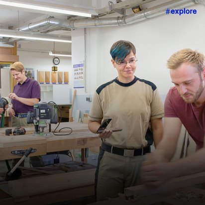 Holzwerkstatt mit mehreren Personen bei der Arbeit; im Vordergrund arbeiten zwei Personen am Holz, während im Hintergrund weitere Personen an Werkbänken mit Werkzeugen hantieren.
