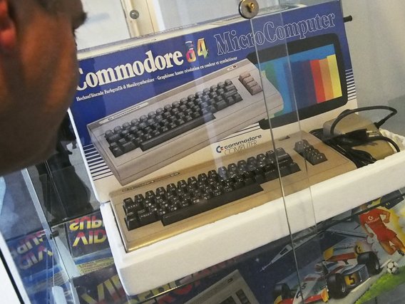 Ein Commodore 64 Mikrocomputer ist ausgestellt, mit seiner charakteristischen beigen Tastatur und bunter Verpackung mit mehrsprachigem Text (einschließlich deutscher Bezeichnungen wie "Farbgrafik & Musiksynthesizer").