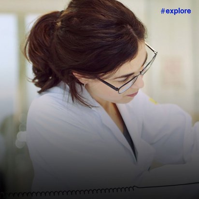 Frau mit Brille in weißem Kittel arbeitet konzentriert an elektronischen Komponenten. Der Hintergrund zeigt ein helles Labor mit unscharfen Lichtpunkten.