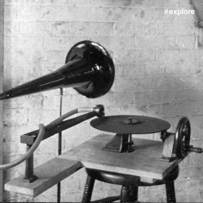 Ein historisches Aufnahmegerät oder früher Phonograph mit trichterförmigem Lautsprecher und Plattenteller auf einem Stuhl montiert, daneben ein großes Rad, vor einer Backsteinwand.