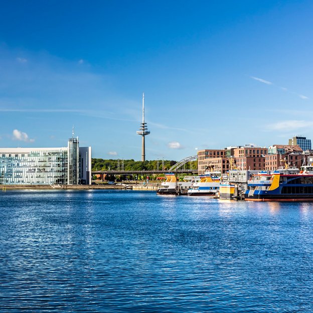 Blick auf den Hafen von Kiel mit modernen Gebäuden und einem Fernsehturm im Hintergrund