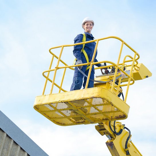Eine Person steht in einer gelben Hubarbeitsbühne, die neben einem Gebäude positioniert ist. Sie trägt einen Schutzhelm und Arbeitskleidung, während sie in der Höhe arbeitet. Der Himmel ist klar und blau, was auf einen sonnigen Tag hinweist.