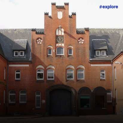Historisches Backsteingebäude mit zwei markanten Spitzdächern und einem zentralen Uhrenturm im Burgstil, umgeben von Bäumen und mit sichtbarer Einfahrt.