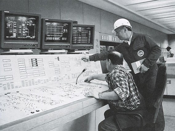 Zwei Techniker in einem Kernkraftwerk-Kontrollraum der 1970er Jahre vor Monitoren und einem ausgedehnten Schaltpult.