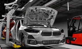 Ein silberner BMW in einer Autofabrik mit geöffneter Motorhaube und teilweise montierter Karosserie auf einer Produktionslinie. Rechts ist ein roter Gabelstapler zu sehen.