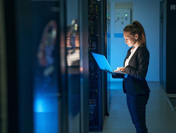 Cybersicherheit in überwachungsbedürftigen Anlagen Ein Frau mit einem Laptop in den Händen steht in einem Rechenzentrum. Sie schaut auf den Bildschirm des Laptops.