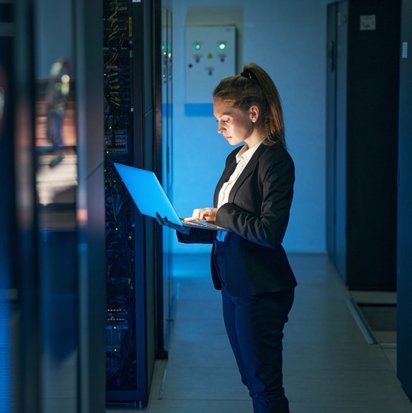 Ein Frau mit einem Laptop in den Händen steht in einem Rechenzentrum. Sie schaut auf den Bildschirm des Laptops.