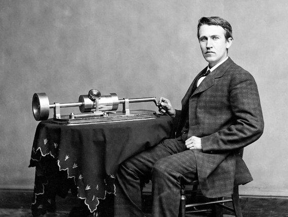 Thomas Alva Edison im formellen Anzug sitzt an einem tischartigen Gerät mit mechanischen Komponenten, vermutlich ein frühes Tonaufnahmegerät oder Phonograph. Der Hintergrund ist neutral und grau.