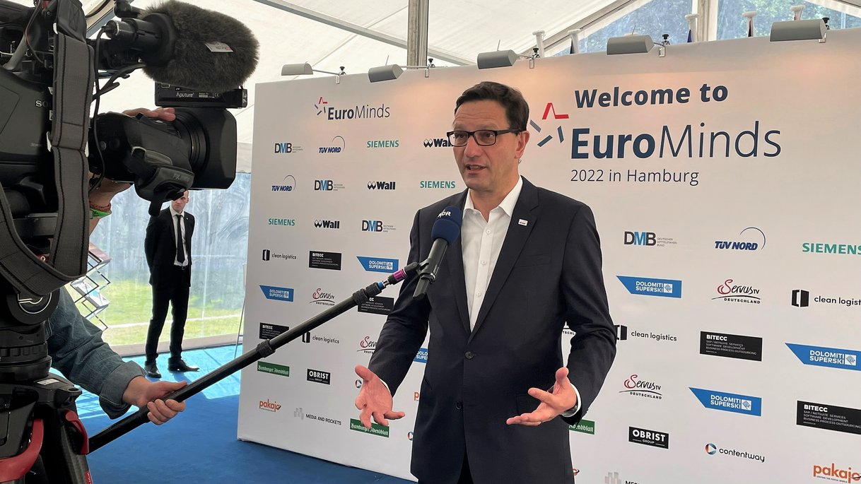Ein Mann im Anzug gibt ein Interview vor einer Kamera mit "Welcome to EuroMinds 2022 in Hamburg"-Hintergrund und Sponsorenlogos.