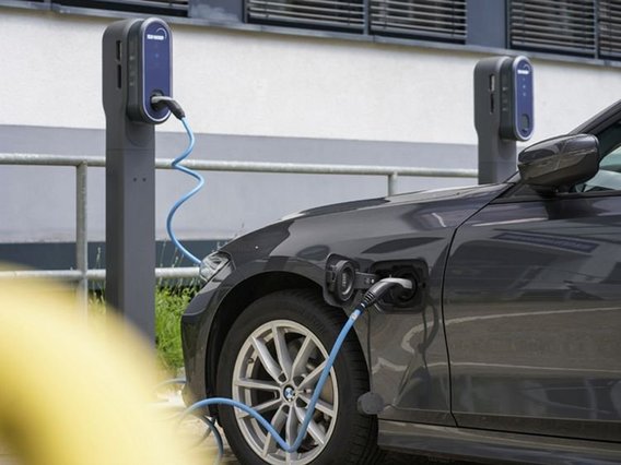 Ein dunkelgraues Elektrofahrzeug lädt an einer öffentlichen Ladestation. Das blaue Kabel verbindet das Auto mit der Station, während im Hintergrund eine Reihe ähnlicher Ladestationen zu sehen ist.