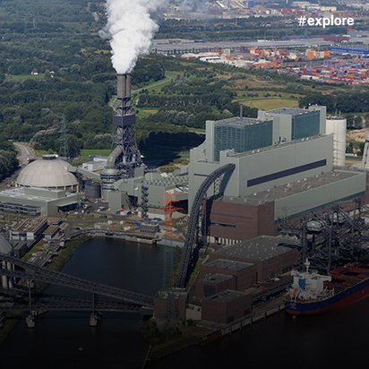 Luftaufnahme eines Industriekomplexes am Wasser mit Kraftwerk, Kühltürmen und Hafenanlagen, erkennbar an den rauchenden Schornsteinen und Containerterminals im Hintergrund.
