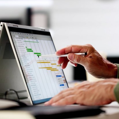 Hand zeigt mit Stift auf ein Gantt-Diagramm zur Projektplanung auf einem Laptop-Bildschirm.