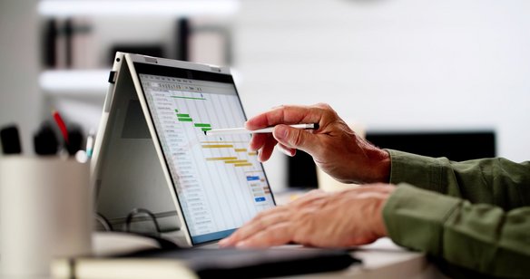 Hand zeigt mit Stift auf ein Gantt-Diagramm zur Projektplanung auf einem Laptop-Bildschirm.