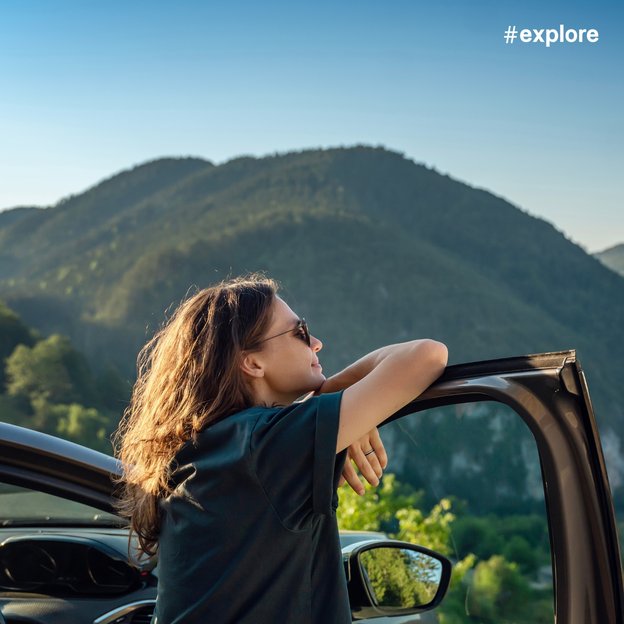 Eine Person lehnt sich entspannt an die geöffnete Autotür und blickt auf eine malerische Berglandschaft. Die Sonne scheint hell und die grünen Hügel erstrecken sich bis zum Horizont.
