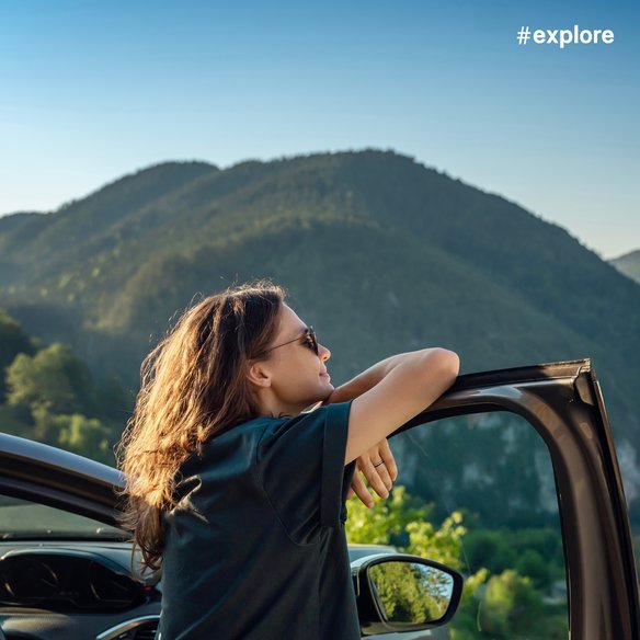 Eine Person lehnt sich entspannt an die geöffnete Autotür und blickt auf eine malerische Berglandschaft. Die Sonne scheint hell und die grünen Hügel erstrecken sich bis zum Horizont.