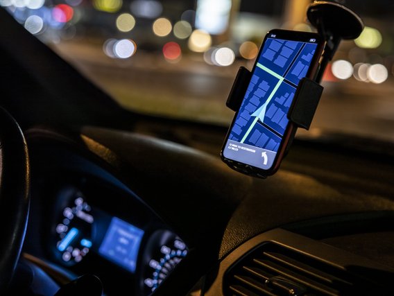 Ein Smartphone in einer Autohalterung zeigt eine blaue Navigationskarte, im Hintergrund sind verschwommene Stadtlichter zu sehen. Das Gerät dient als GPS-Navigation im Fahrzeuginneren bei nächtlicher Fahrt.