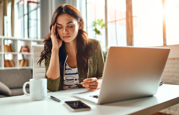 Am Arbeitsplatz mit Laptop, Kaffee und Smartphone wirkt eine Frau erschöpft und nachdenklich – ein Moment mentaler Belastung im Büroalltag.