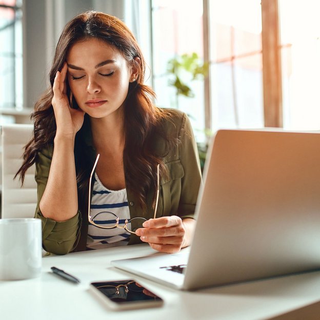 Am Arbeitsplatz mit Laptop, Kaffee und Smartphone wirkt eine Frau erschöpft und nachdenklich – ein Moment mentaler Belastung im Büroalltag.