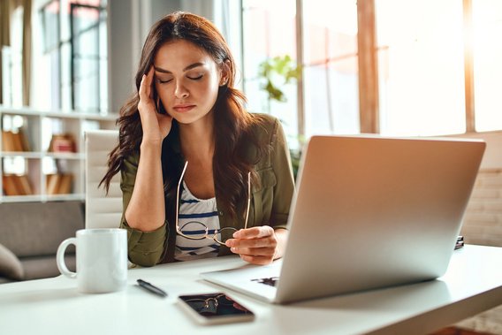 Am Arbeitsplatz mit Laptop, Kaffee und Smartphone wirkt eine Frau erschöpft und nachdenklich – ein Moment mentaler Belastung im Büroalltag.
