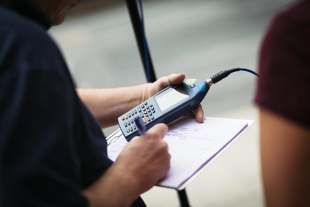 Detailaufnahme einer Hand, die ein Schallpegelmessgerät über einem Formular hält, verbunden mit einem Audiokabel.