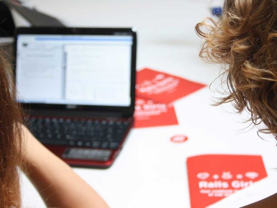 Zwei Frauen mit braunem Haar arbeiten gemeinsam an einem Laptop, im Hintergrund sind rote Dokumente oder Materialien sichtbar.