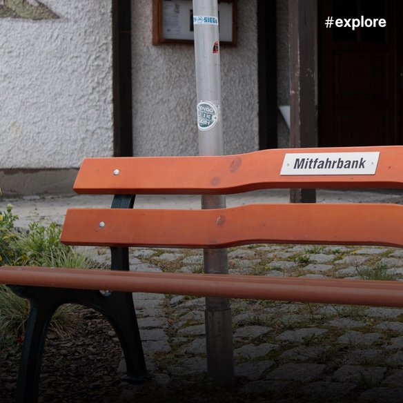Orange Parkbank mit Schild "Mitfahrbank" vor einem traditionellen Fachwerkhaus auf einem Kopfsteinpflasterplatz.