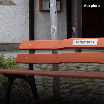 Orange Parkbank mit Schild "Mitfahrbank" vor einem traditionellen Fachwerkhaus auf einem Kopfsteinpflasterplatz.