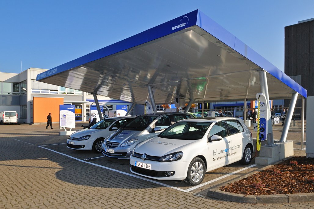  Unter einem blauen Überdach mit der Aufschrift "TÜV NORD STATION" stehen mehrere Autos geparkt. Im Vordergrund ist ein weißer Volkswagen mit der Aufschrift "blue-e-motion Der elektrische Volkswagen" zu sehen. Die Fahrzeuge sind auf einem gepflasterten Parkplatz abgestellt, und im Hintergrund sind moderne Gebäude und Informationsstände zu erkennen.