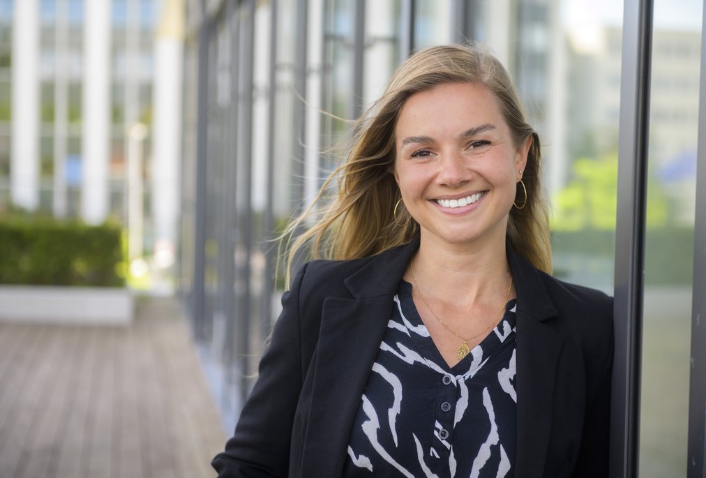 Eine Frau mit langen, blonden Haaren steht in einem modernen städtischen Umfeld neben einer Glasfassade. Sie trägt einen schwarzen Blazer und ein gemustertes Oberteil. Im Hintergrund sind unscharf Gebäude und Grünflächen zu erkennen.