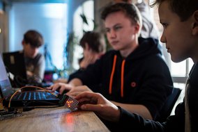 Jugendliche an Laptops mit einer LED-Matrix, die rötlich leuchtet, in einem hellen Arbeitsraum.