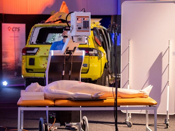 Crashtest-Dummy auf einer Untersuchungsliege mit Messgerät, dahinter ein beschädigtes gelbes Testfahrzeug.
